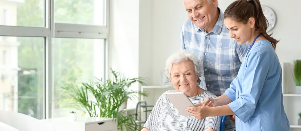 Pflegekraft zeigt älterer Patientin und Angehörigem digitale Pflegeinformationen auf einem Tablet.